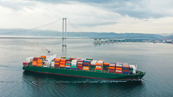 Drone shot of large cargo ship carrying containers passing under the bridge